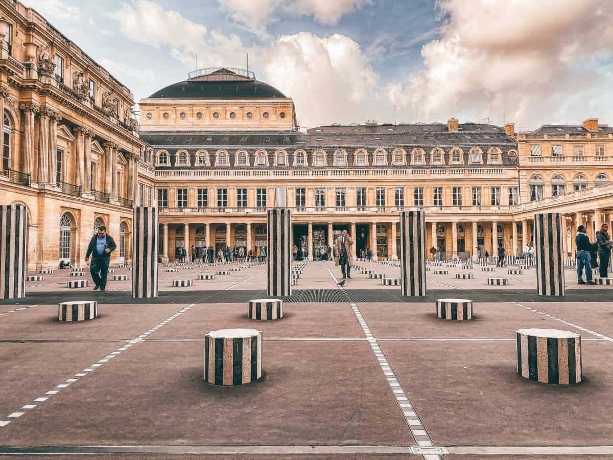 People walking around a court yard filled
with tall and short black and white columns in front of an old building