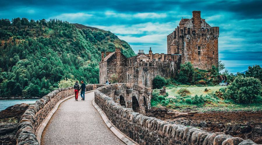 A couple crossing the bridge to The ruin of a castle sitting on the side of a lake