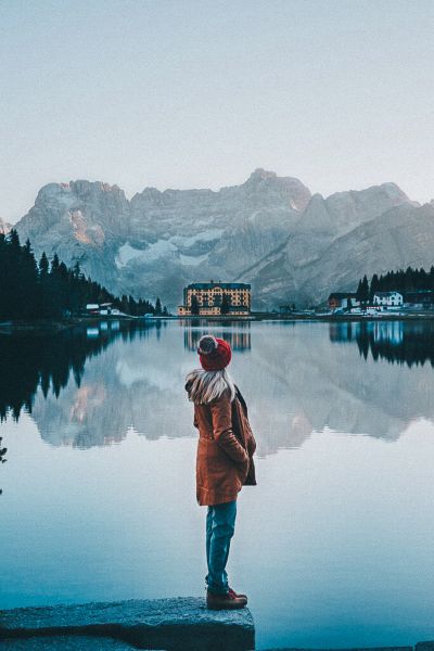 A blonde girl wearing a red beanie standing on the edge of Lake Misurina
