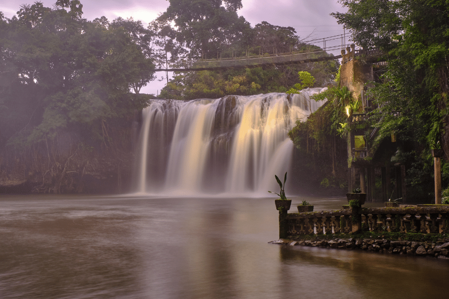 A rope bridge crossing Mena Creek Falls with a ruined building to the right of the falls