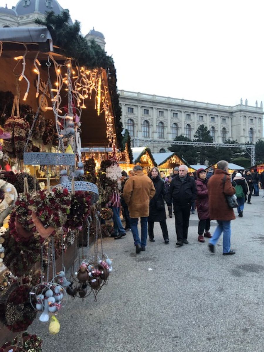 Visitors milling around a festive Christmas market in 3 days in Vienna at Christmas, with a decorated stall in the foreground showcasing wreaths and ornaments, and the elegant architecture of a grand building in the background