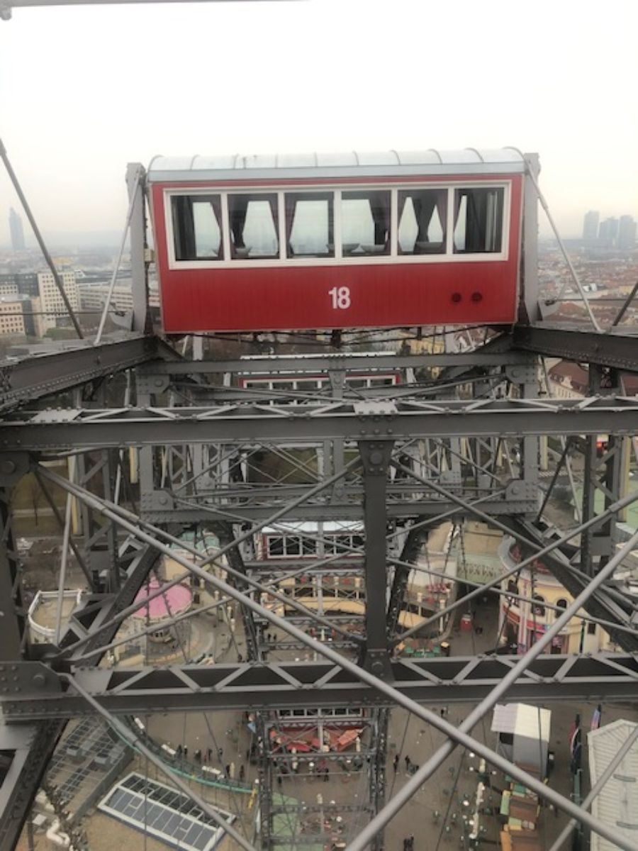 A red cabin number 18 of a Ferris wheel, poised high above ground within a complex metal structure, with an aerial view of the cityscape and smaller structures below