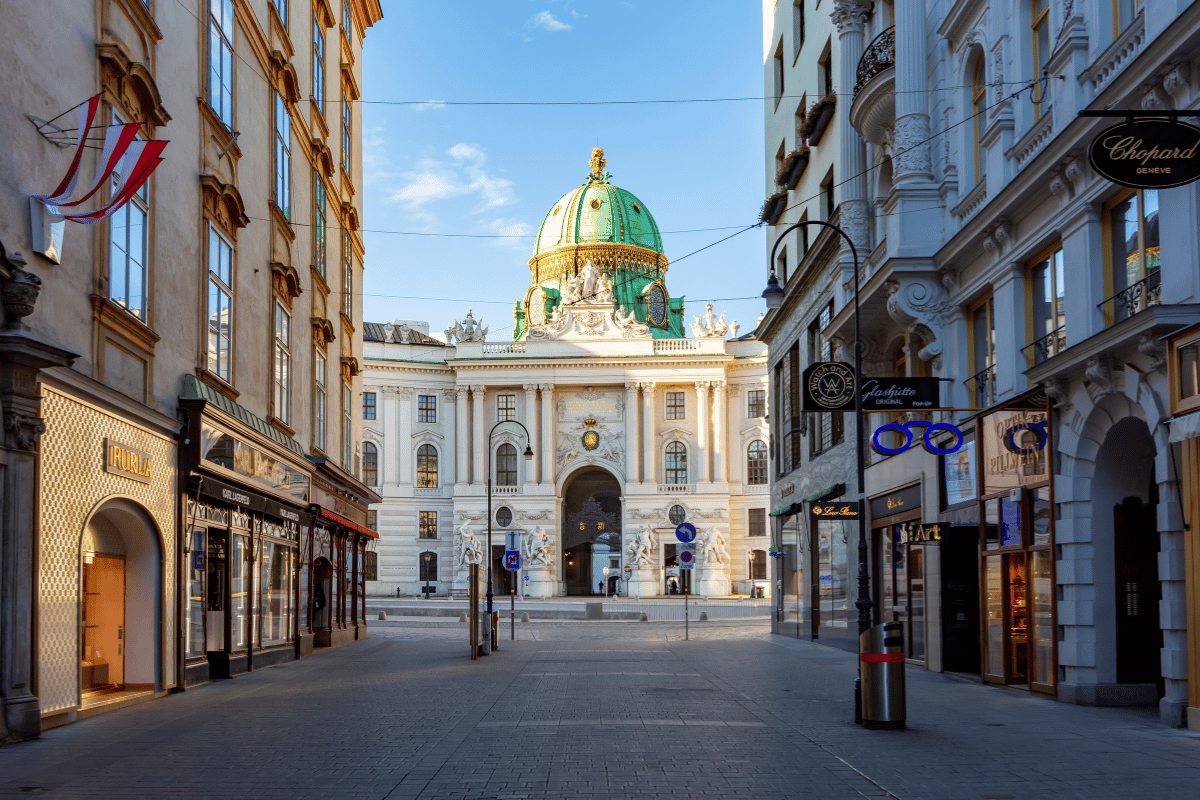 A serene morning on Kohlmarkt street in Vienna, leading to the richly adorned Hofburg Palace with its distinctive green dome. The street is lined with high-end shops and a few pedestrians, framed by the warm glow of the rising sun, creating a tranquil urban scene